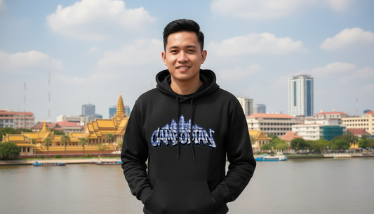 Person wearing a black hoodie with 'Capital of the World' text, standing by a waterfront with buildings in the background.