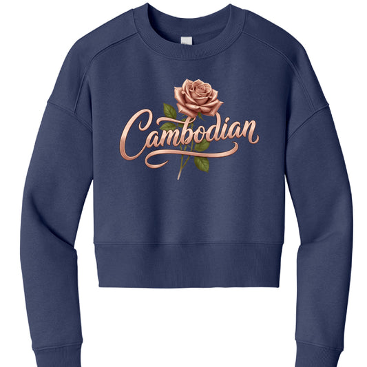 This image features a navy blue, cropped sweatshirt displayed against a plain white background. The garment has a casual, modern fit with a round crew neck and ribbed cuffs and hem.
The central focus of the sweatshirt is a graphic design that includes:
Floral Illustration: A realistic, rose-gold or dusty pink rose in full bloom, complete with green leaves and a stem.
Typography: The word "Cambodian" is written across the rose in a stylized, rose-gold metallic script. The font features elegant flourishes,