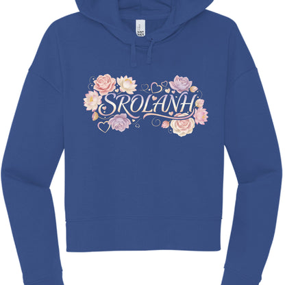 This image features a cobalt blue, cropped hoodie displayed against a plain white background. The garment is laid flat, showcasing a decorative graphic centered on the chest.

Design Details
Text: The word "SROLANH" is written in a stylized, elegant serif font. The "S" and the "H" feature decorative flourishes that sweep underneath the rest of the letters.

Floral Elements: The text is framed by a cluster of soft-colored flowers, primarily roses and lotus blossoms in shades of pale pink, lavender, and cream