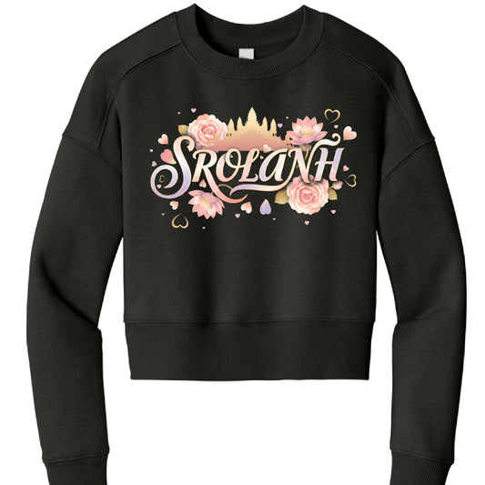 The image features a black, long-sleeved cropped sweatshirt with a vibrant, illustrative graphic centered on the chest. The design blends cultural symbols with a soft, romantic aesthetic.

Graphic Details
The central focus is the word "SROLANH" (a Khmer word for "love") written in an elegant, stylized script with a pastel gradient of pink, purple, and cream.

Background Silhouette: Behind the text is a golden-tan silhouette of Angkor Wat, the famous Cambodian temple complex.

Floral Accents: The text is sur